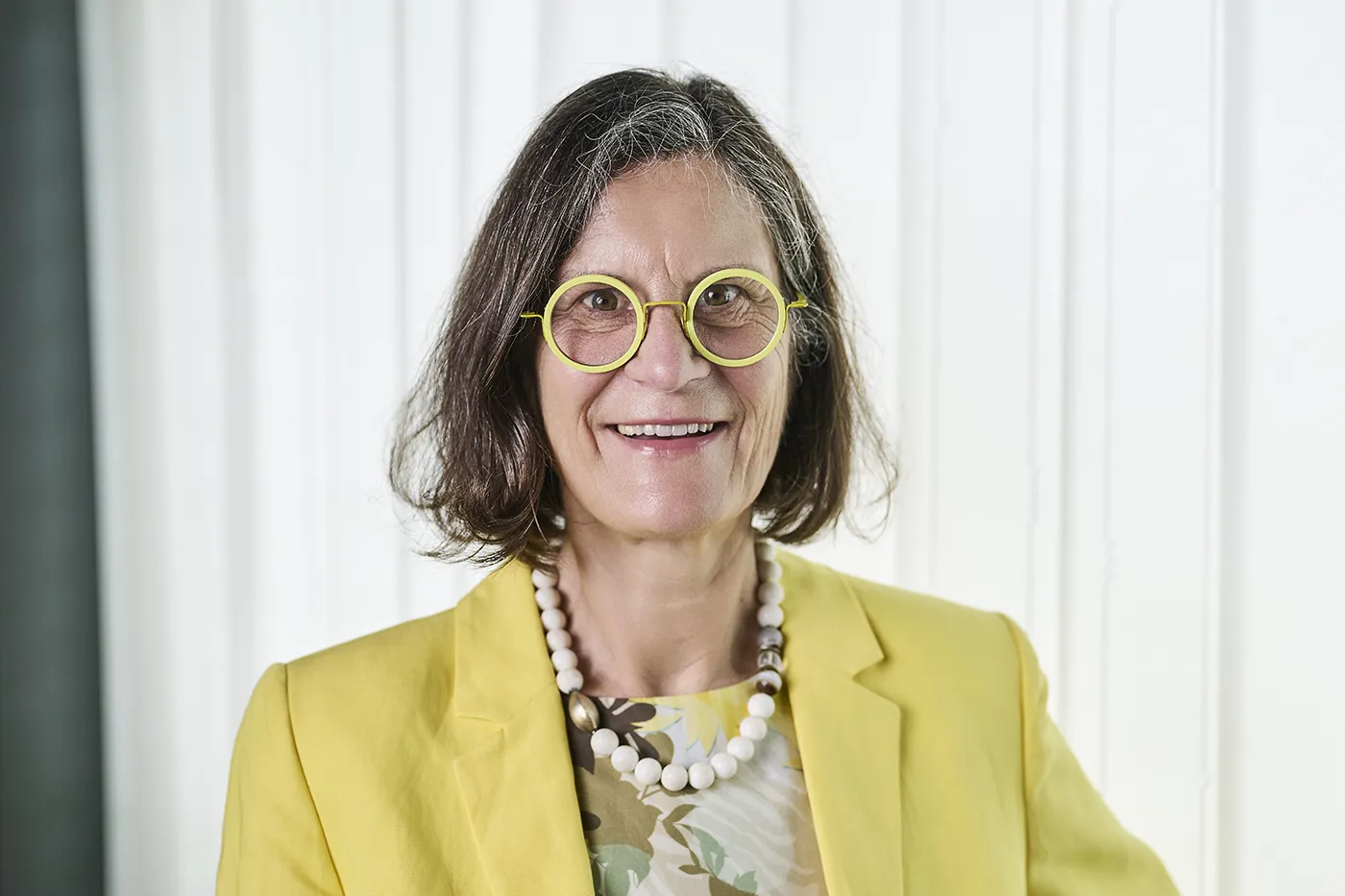 Eine Frau mit schulterlangem braunem Haar, einer gelben Brille und einem gelben Blazer lächelt in die Kamera. Sie trägt ein geblümtes Oberteil und eine klobige weiße Perlenkette. Im Hintergrund sind weiße Vorhänge zu sehen.
